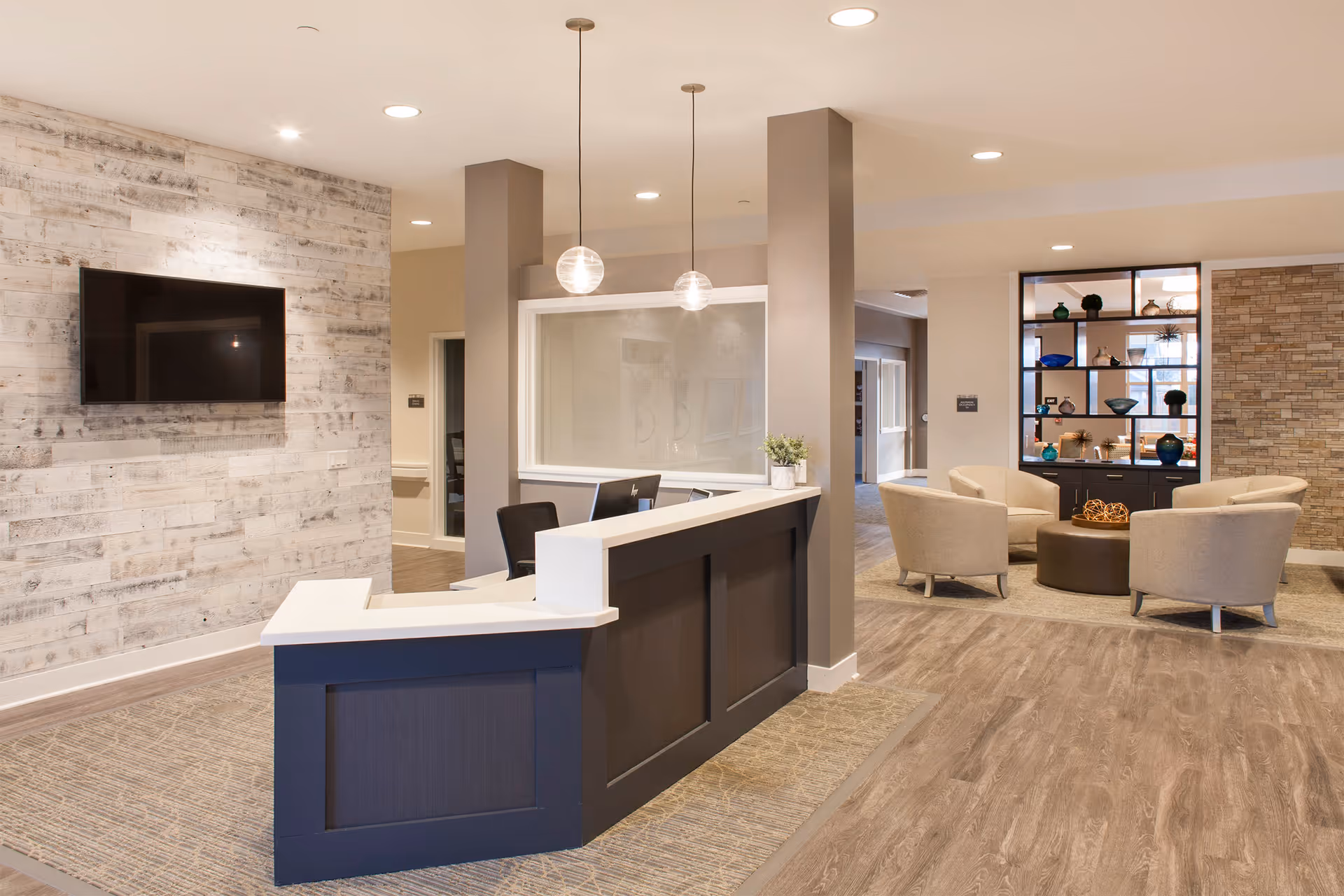 Modern reception desk and comfortable seating area in a senior living facility lobby with a wall-mounted TV and decorative shelving.