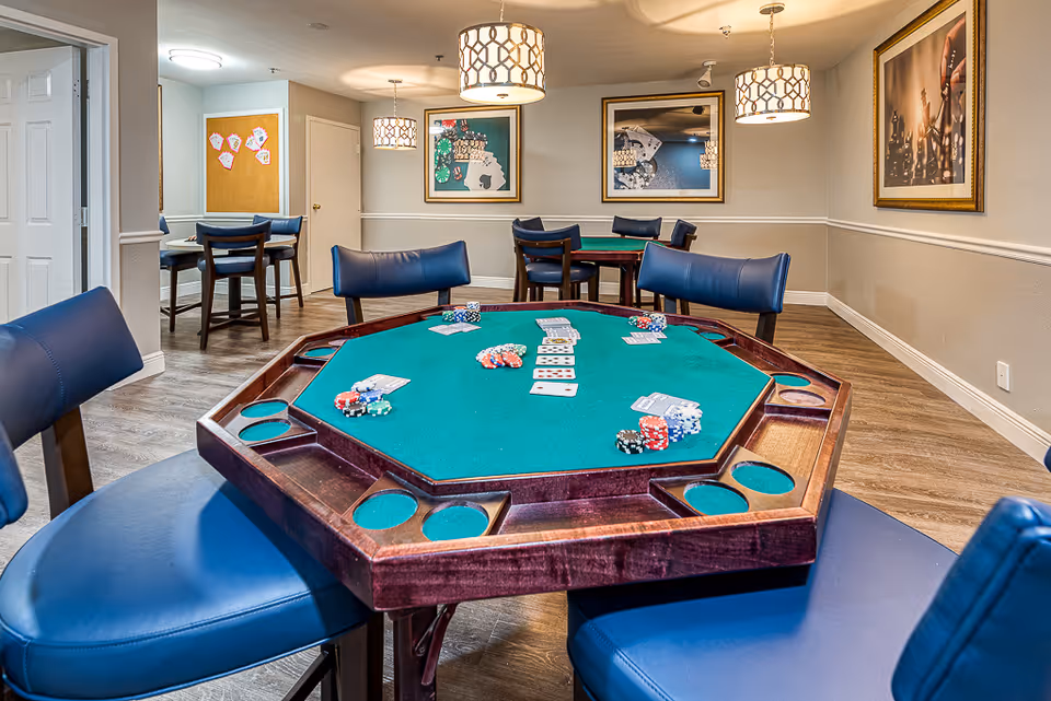 Interior view of a game room with a green felt octagonal poker table surrounded by blue cushioned chairs. The table has poker chips and playing cards on it. The room has wooden flooring, beige walls with white trim, framed pictures on the walls, and three hanging pendant lights. In the background, there is another table with chairs and a bulletin board on the wall.