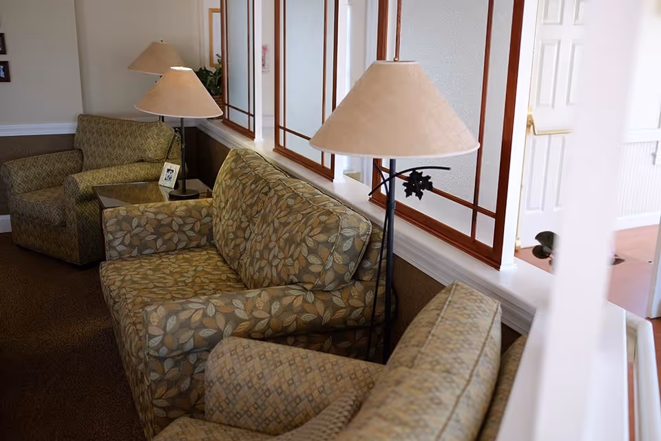 A cozy seating area with patterned upholstered armchairs and a loveseat, accompanied by two floor lamps with beige lampshades. The space is softly lit and features frosted glass windows with wooden frames and a white door in the background.