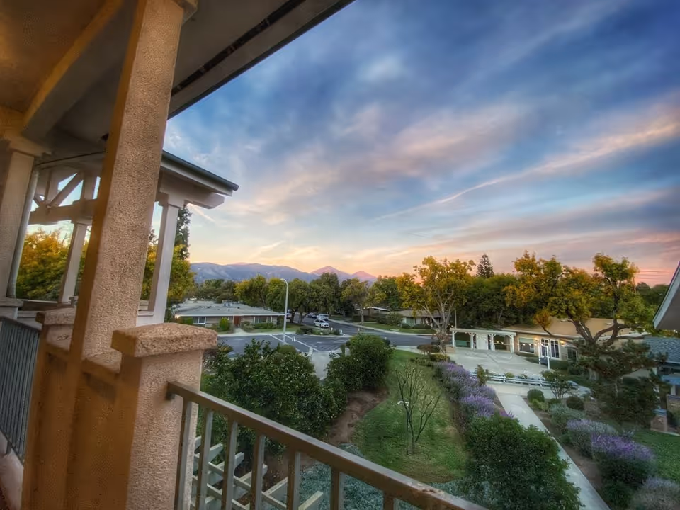 View from a covered balcony over a landscaped courtyard and single-story buildings with trees and distant mountains under a colorful sunset sky.