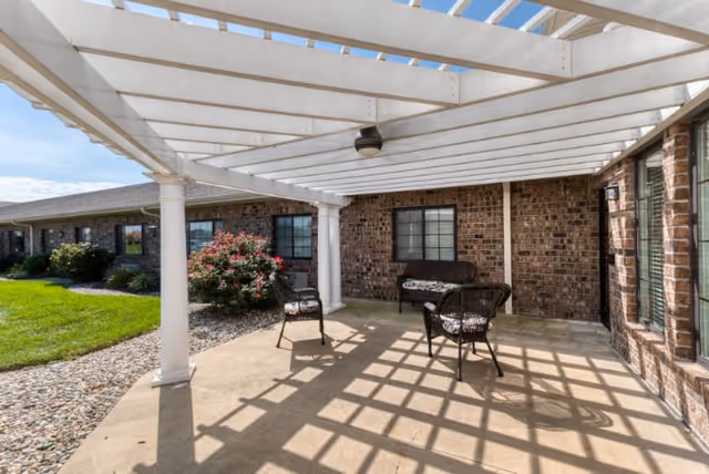 Outdoor patio area with a white pergola casting shadows on the concrete floor. There are three chairs and a bench with cushions arranged under the pergola. The patio is adjacent to a brick building with windows and surrounded by a landscaped area with grass and bushes.