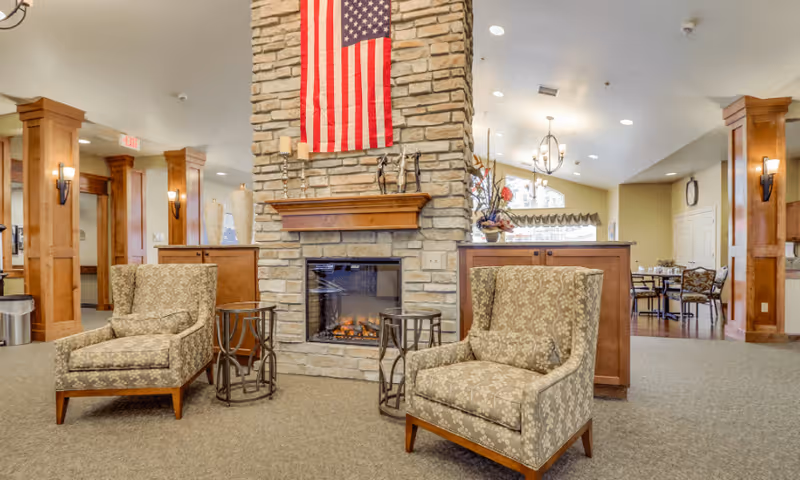 A cozy common area in an assisted living facility featuring two patterned armchairs facing a stone fireplace with an American flag hanging above it. The room has wooden columns, soft lighting, and a dining area visible in the background with tables and chairs.
