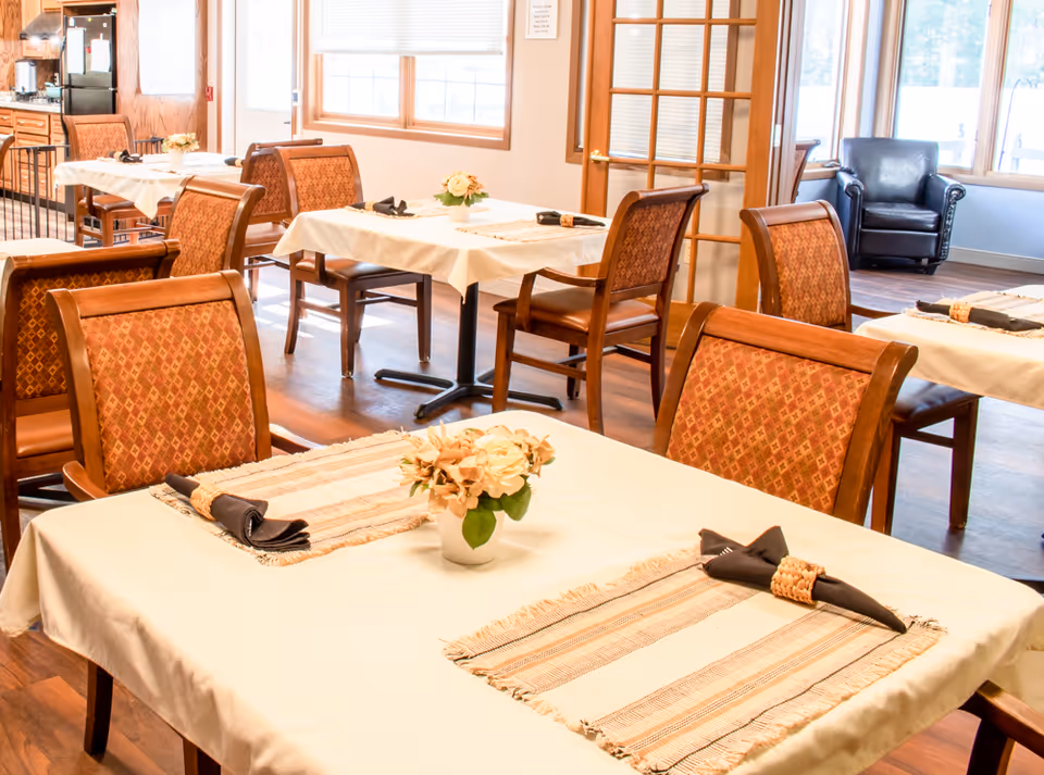 Bright dining room with neatly set tables, upholstered chairs, and floral centerpieces.