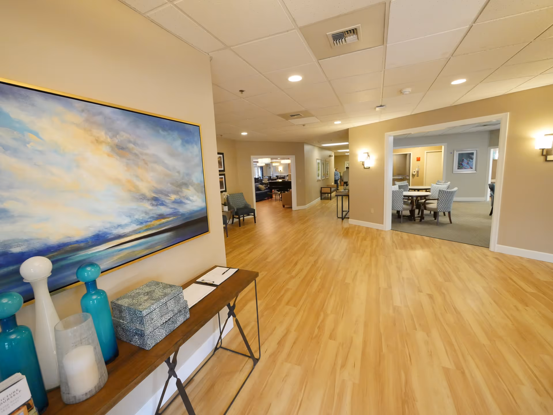 Interior view of a senior living facility hallway with light wood flooring and beige walls. On the left side, there is a console table with decorative blue and white vases, a candle, and a large abstract painting above it. The hallway leads to a room with a round table and chairs, and further back, a lounge area with seating is visible. The ceiling has recessed lighting and air vents.