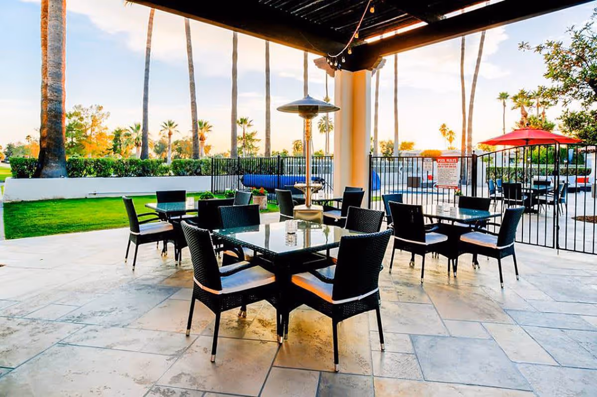 Outdoor covered patio area with several black wicker tables and chairs, a tall outdoor heater, and a view of palm trees and green grass beyond a black metal fence.