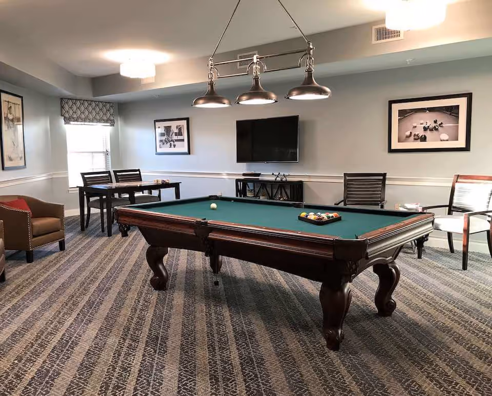 A recreational room with a pool table in the center, a set of billiard balls arranged on the table, and a single cue ball. The room has patterned carpet flooring, light blue walls with white trim, and three framed pictures on the walls. There is a flat-screen TV mounted on the wall above a small console table. To the left, there is a small table with two chairs near a window with a patterned valance. Additional seating includes a brown armchair with a red cushion and two other chairs with armrests.