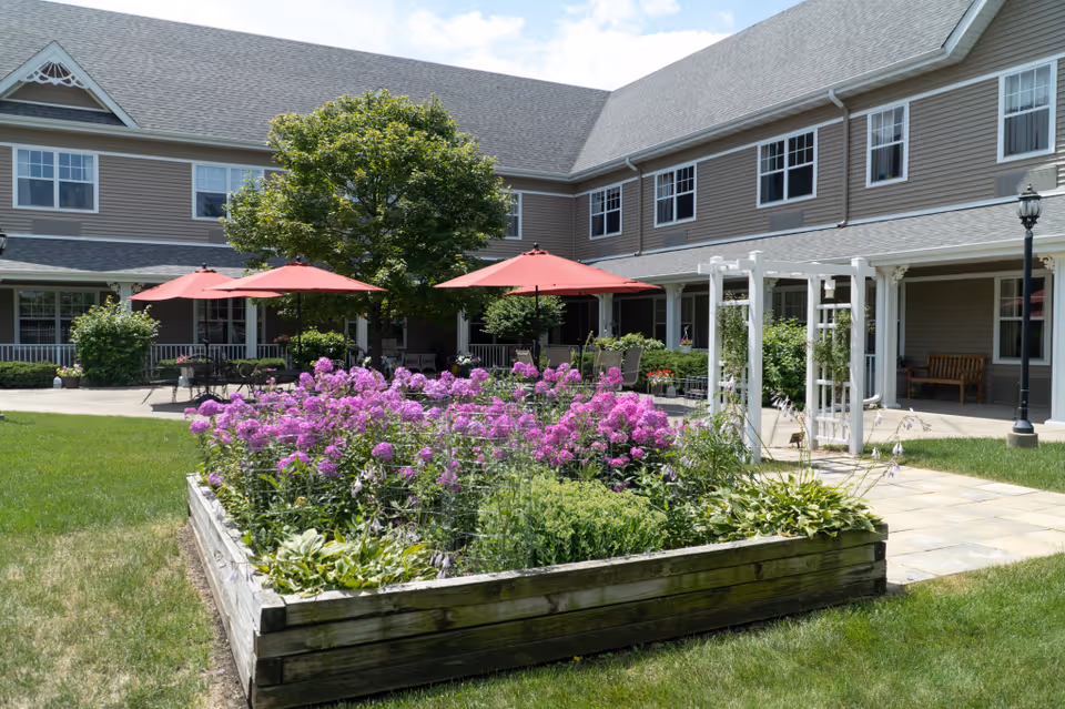 Outdoor garden area at Grand Victorian of Rockford featuring a raised wooden flower bed with blooming purple flowers, green shrubs, and a white trellis. In the background, there are several red patio umbrellas shading tables and chairs on a paved patio, with a two-story building with multiple windows surrounding the courtyard.