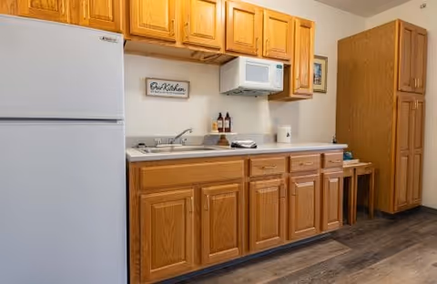 A small kitchen area with wooden cabinets, a white refrigerator, a white microwave mounted above the counter, a stainless steel sink, and a few decorative items including a framed sign that says 'Our Kitchen'. The floor is wood-patterned and there is a tall wooden pantry cabinet on the right side.