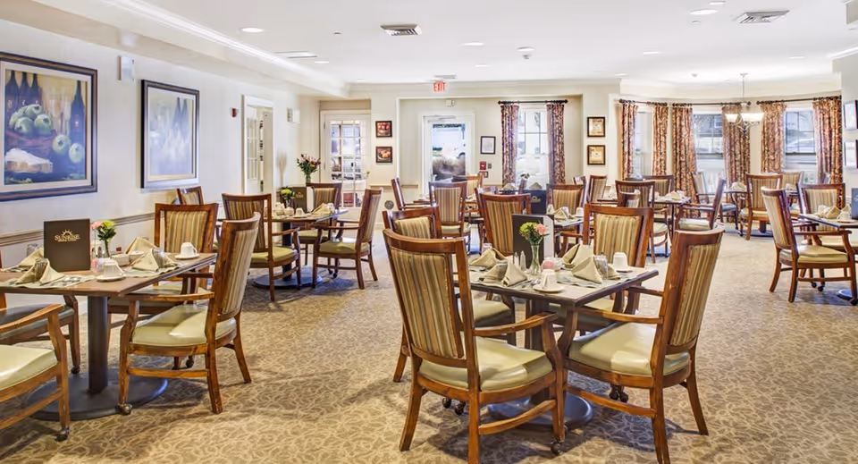A bright and spacious dining room in a senior living facility with multiple wooden tables and chairs arranged neatly. Each table is set with napkins, cups, and small flower vases. The room has patterned carpet flooring, framed artwork on the walls, and large windows with floral curtains allowing natural light to enter.