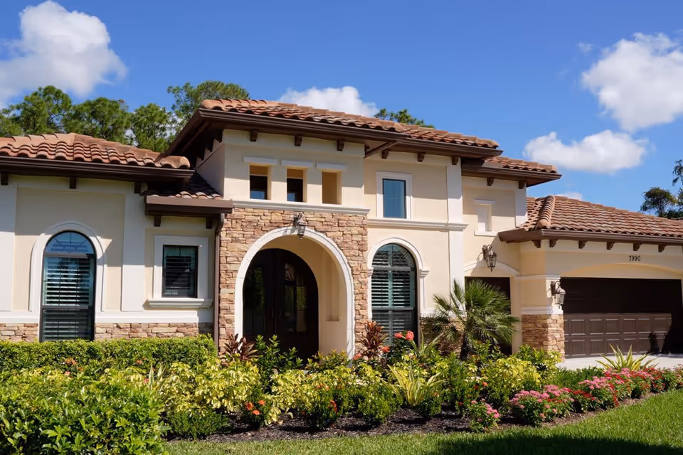 Exterior view of a single-story residential building with a tiled roof, arched front entrance, stone and stucco facade, surrounded by well-maintained landscaping including bushes and flowering plants under a blue sky with some clouds.