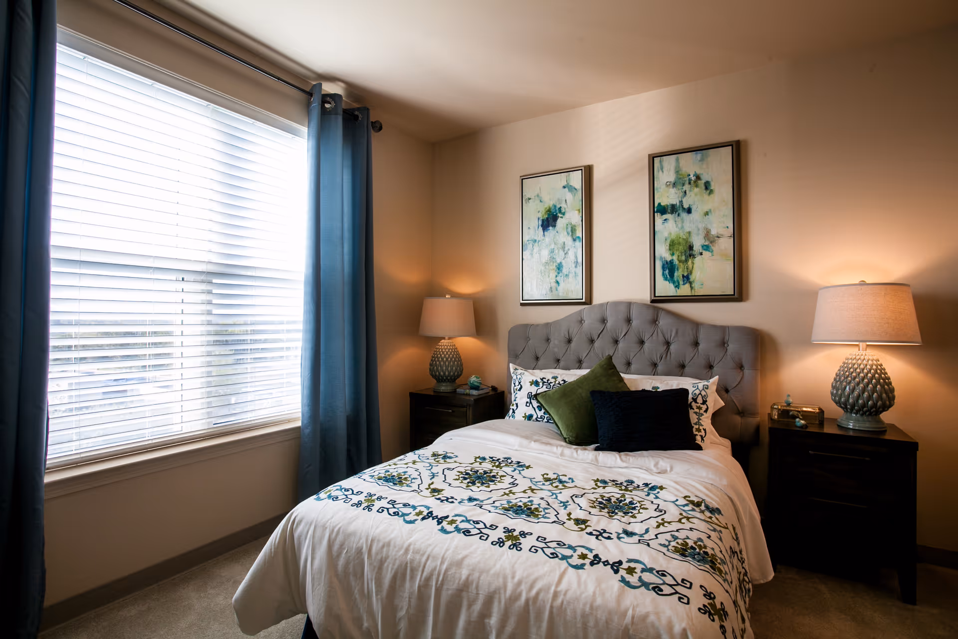 A neatly made bedroom with a tufted headboard, patterned bedding, two bedside tables with lamps, framed artwork, and a large curtained window.