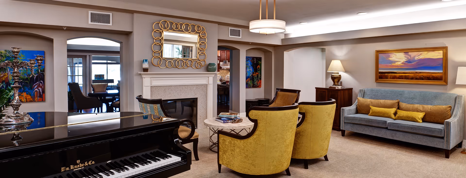 A cozy living room area in The Piper Assisted Living and Memory Care featuring a black grand piano in the foreground, two yellow upholstered chairs around a round marble coffee table, a gray sofa with yellow and brown cushions, a fireplace with a decorative mirror above it, and colorful artwork on the walls. The room is warmly lit with a ceiling light and table lamps, and an adjacent room with a dining table and chairs is visible through an open doorway.