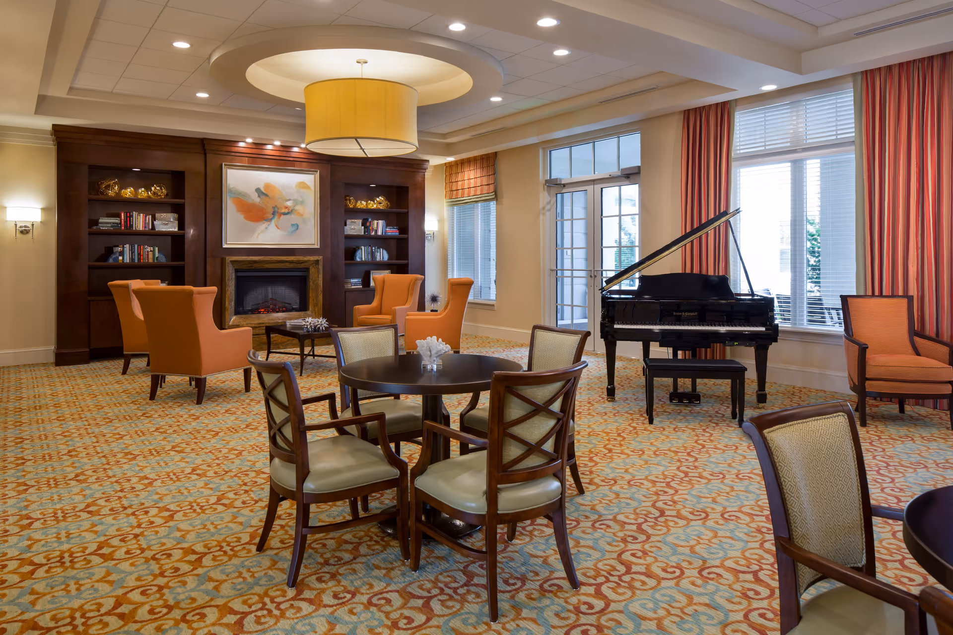 A cozy and elegant common area with a patterned carpet, round tables with chairs, orange armchairs, a grand piano near large windows with red curtains, and a fireplace with built-in bookshelves and decorative items.