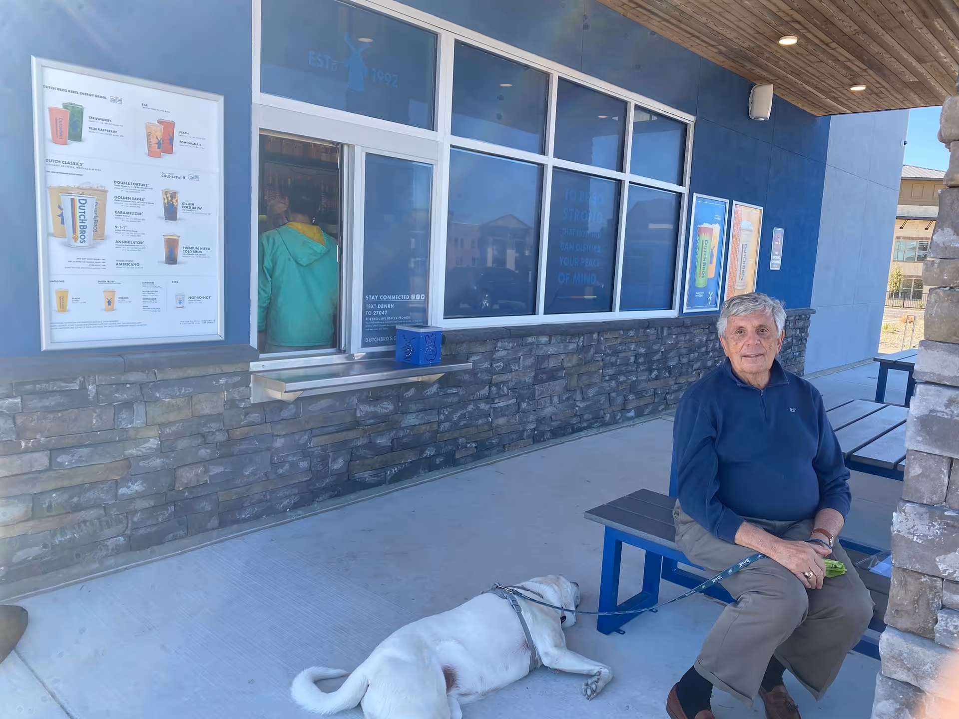 An elderly man sitting on a blue bench outside a building with stone and blue walls. A white dog is lying on the ground next to him. There is a service window with a person inside wearing a green jacket. Menus and advertisements are displayed on the wall near the window.