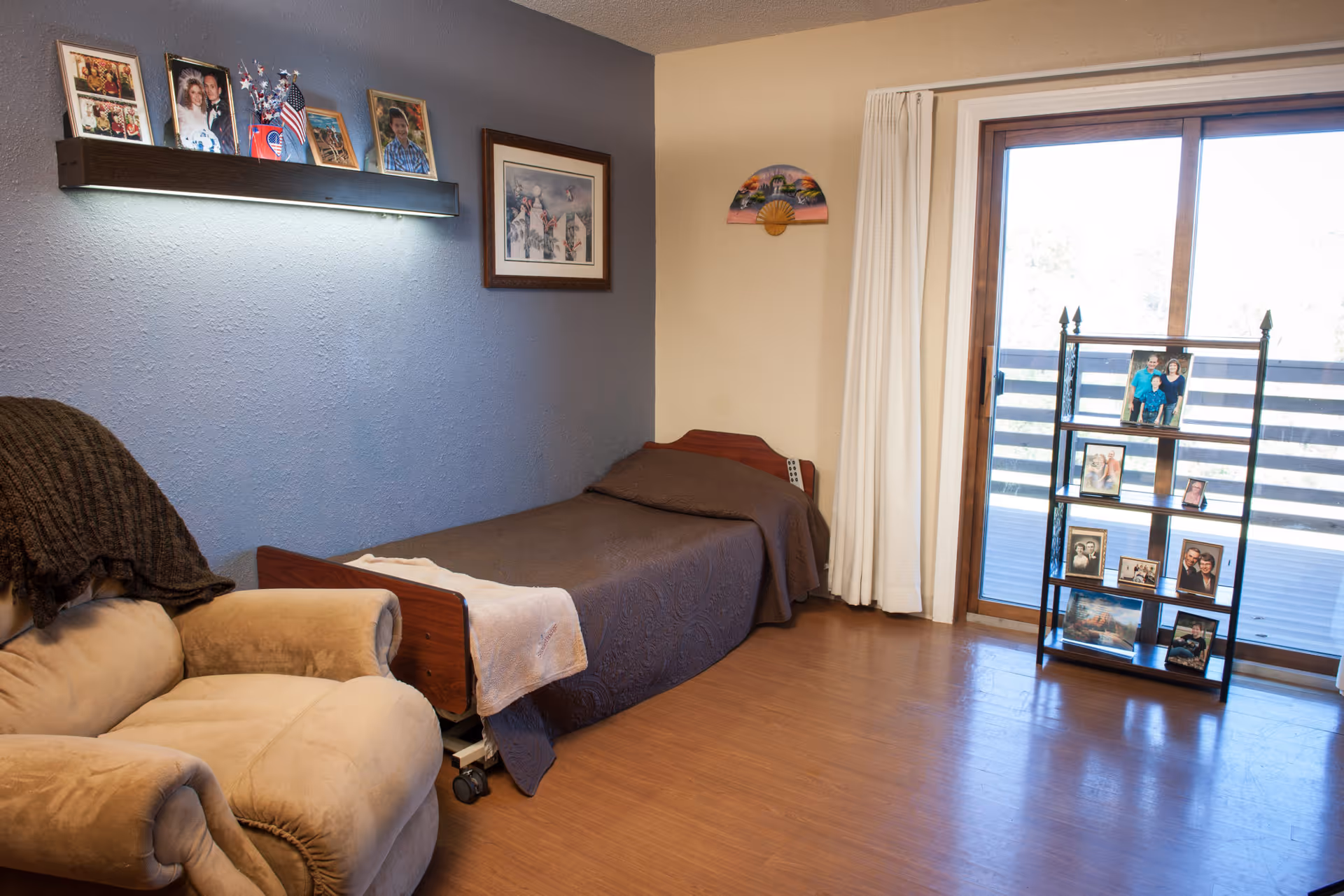 A tidy senior living bedroom with a single bed against a blue wall, an armchair, framed photos on a wall shelf, and a sliding glass door with a small shelving unit of photos.