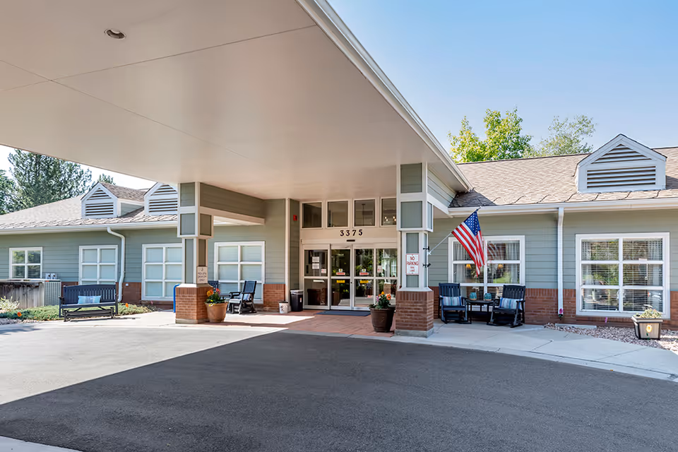 Entrance of Brookdale Boulder Creek facility showing a covered drop-off area with seating benches on both sides, potted plants, an American flag, and large glass doors under the address number 3375.
