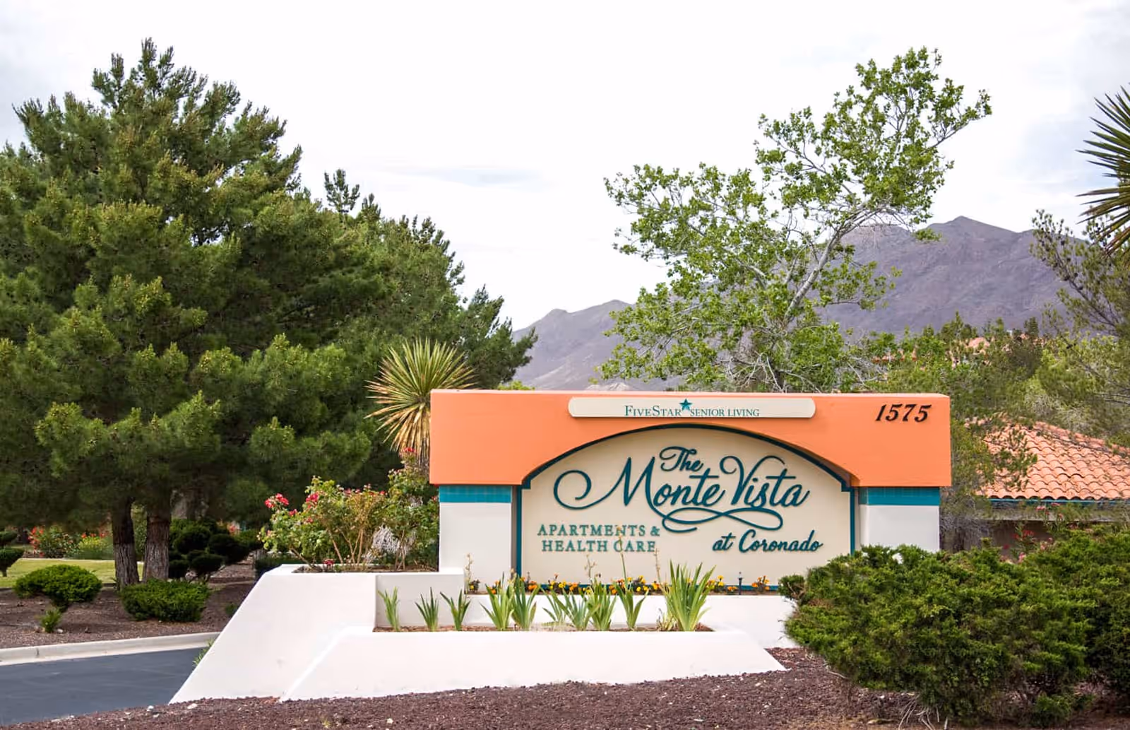Outdoor view of the entrance sign for The MonteVista at Coronado Assisted & Senior Living, surrounded by greenery, trees, and mountains in the background.