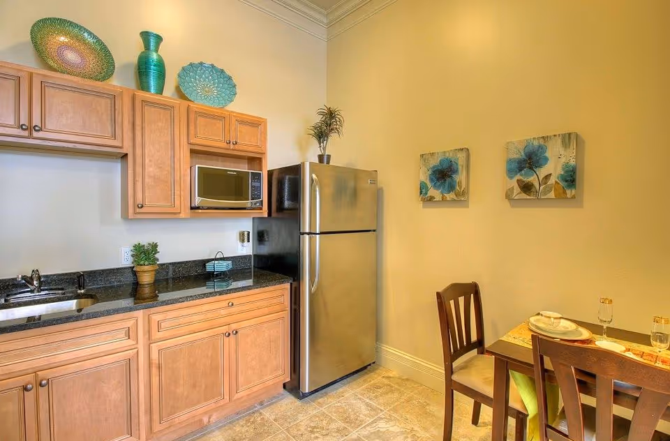 A small kitchen area with wooden cabinets, a stainless steel refrigerator, a microwave, and a sink with a black countertop. There are decorative plates and a vase on top of the cabinets. To the right, there is a dining table set for two with wooden chairs and floral wall art above.
