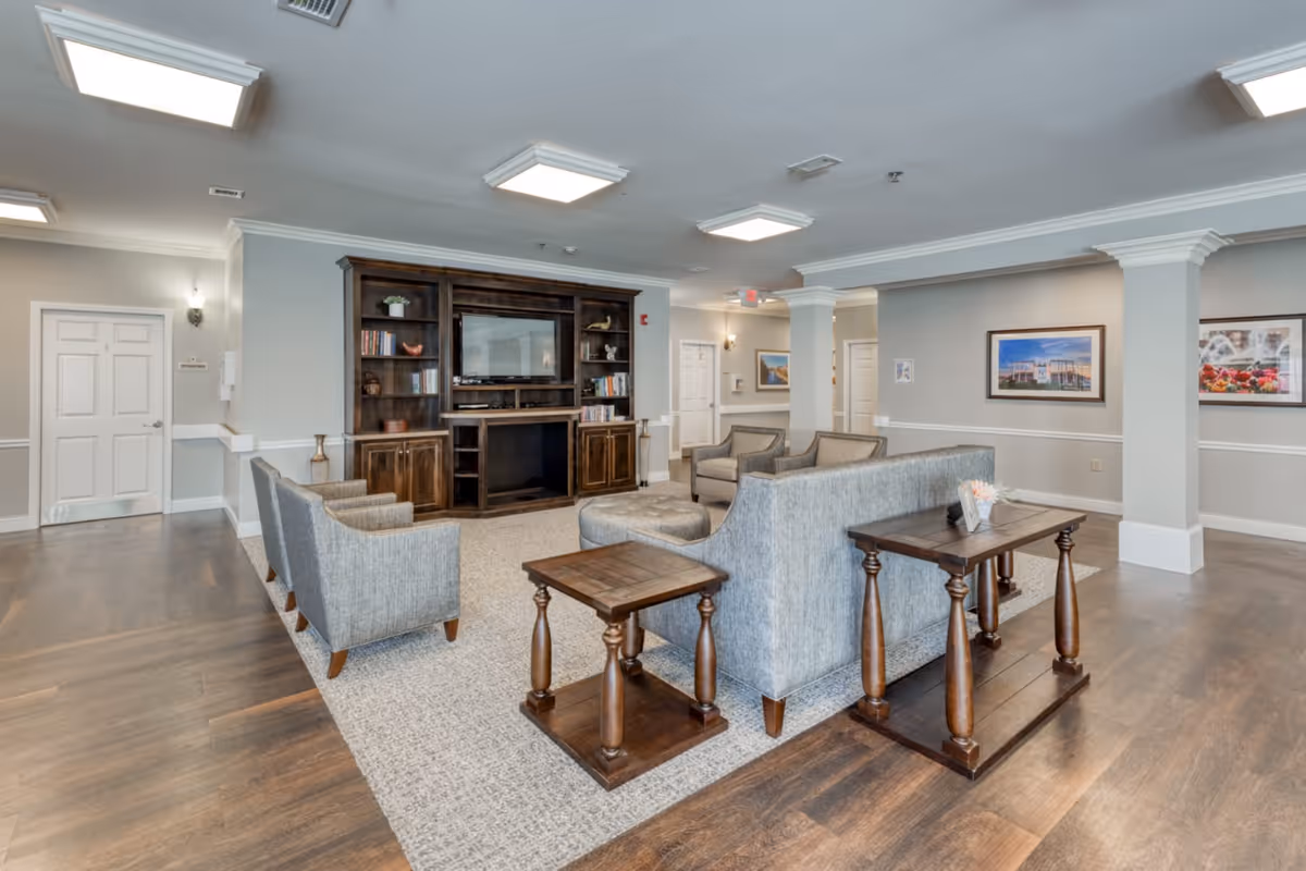 A spacious and well-lit living room area in a senior living facility featuring a large wooden entertainment center with a TV, several gray upholstered chairs and sofas arranged around a central ottoman, wooden side tables, and framed artwork on the walls. The room has hardwood flooring with a large area rug and white crown molding and columns.