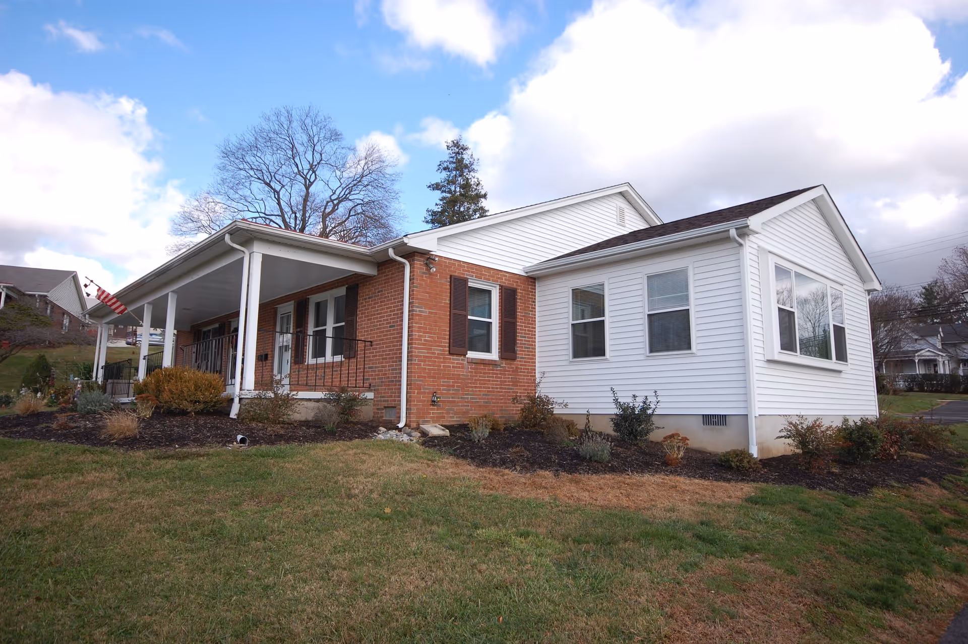 Single-story brick and white-siding house with a covered front porch, lawn and landscaping under a partly cloudy sky.