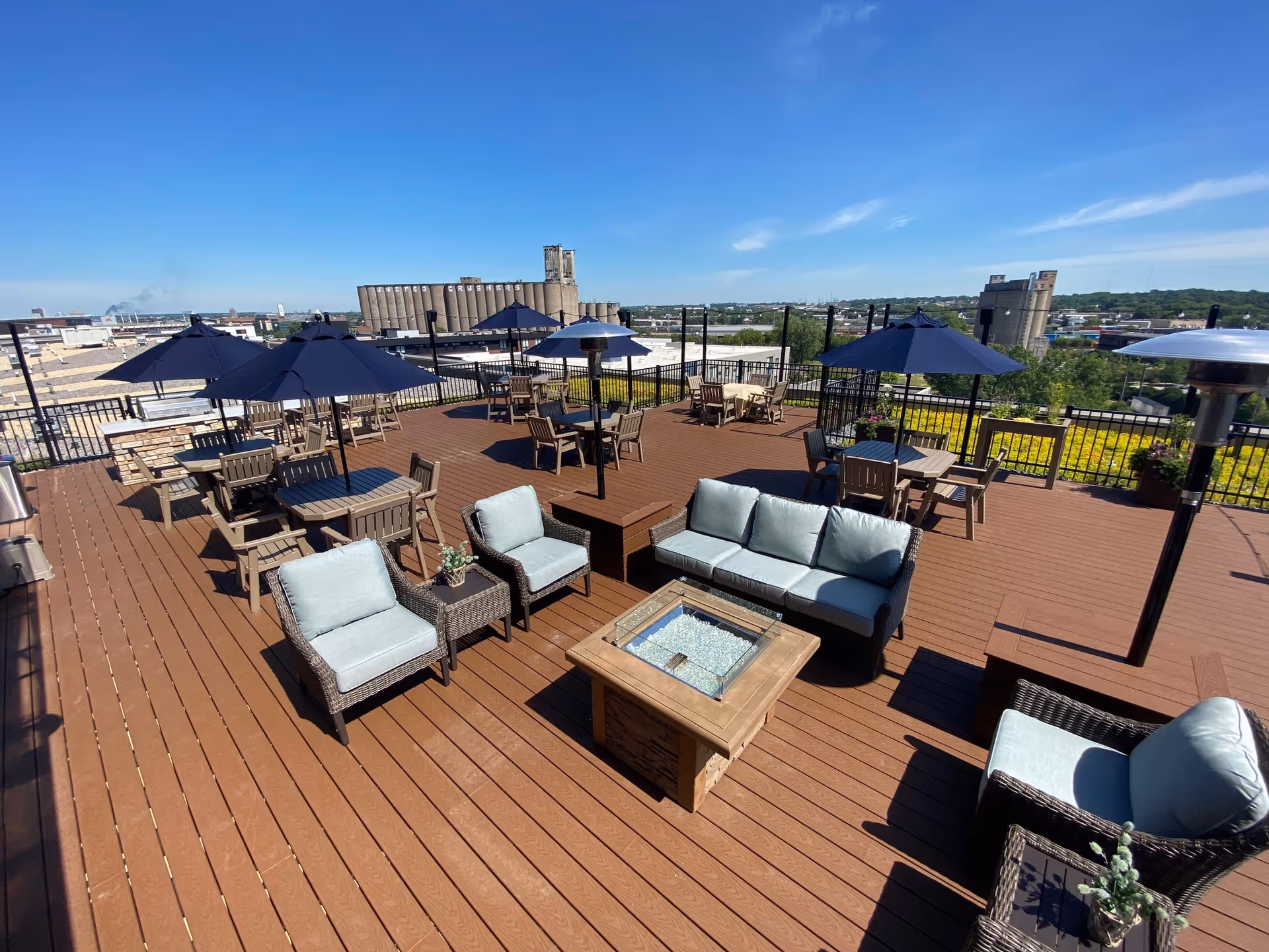 A rooftop patio with wooden decking featuring multiple seating areas including cushioned chairs and sofas around a fire pit, several tables with umbrellas, and outdoor heaters. The patio overlooks an urban landscape with industrial buildings and greenery under a clear blue sky.