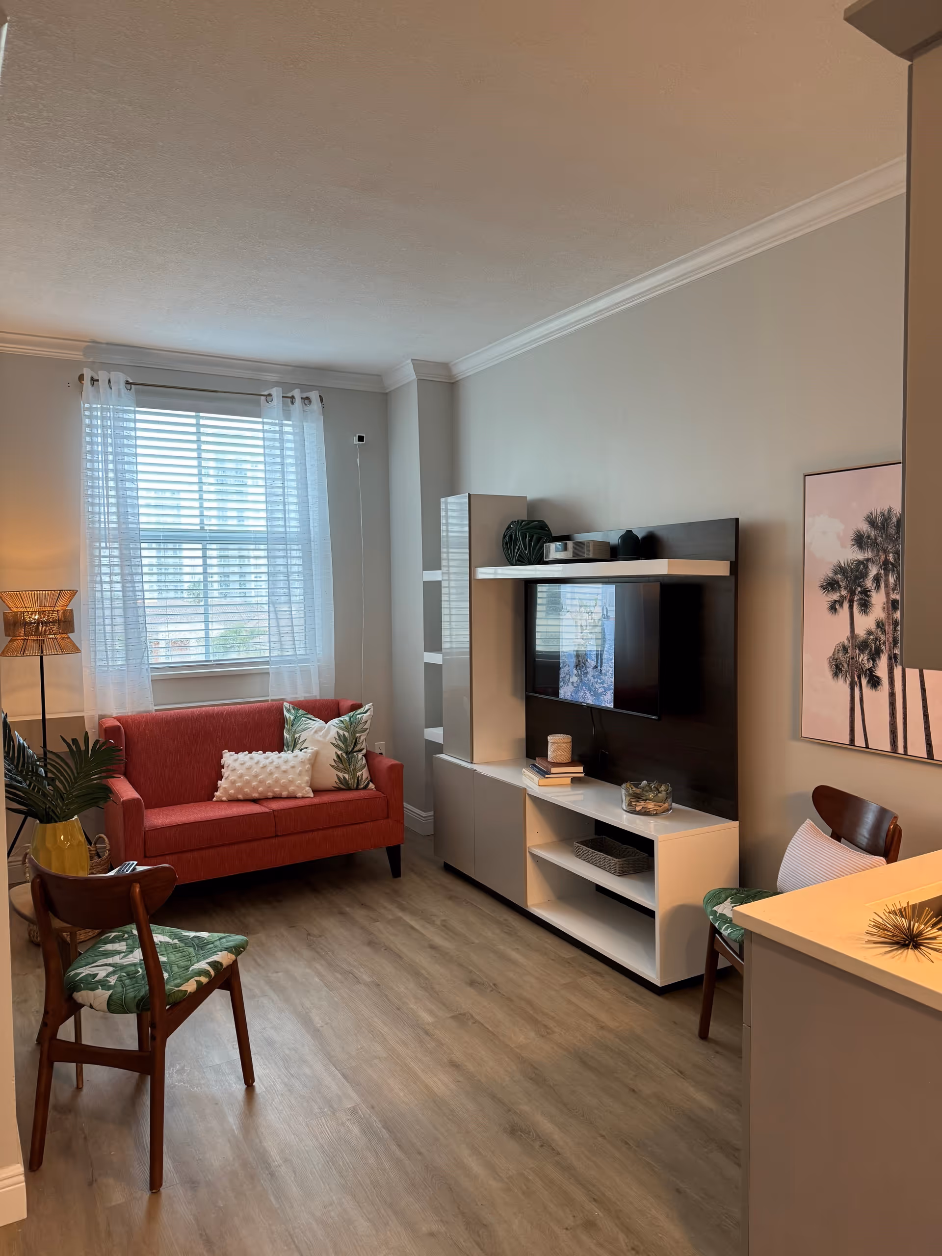 A cozy living room with a red loveseat adorned with two decorative pillows, a wooden chair with a green leaf-patterned cushion, and a modern entertainment unit with a mounted flat-screen TV. The room has light wood flooring, a window with sheer white curtains, a floor lamp, and a framed picture of palm trees on the wall.