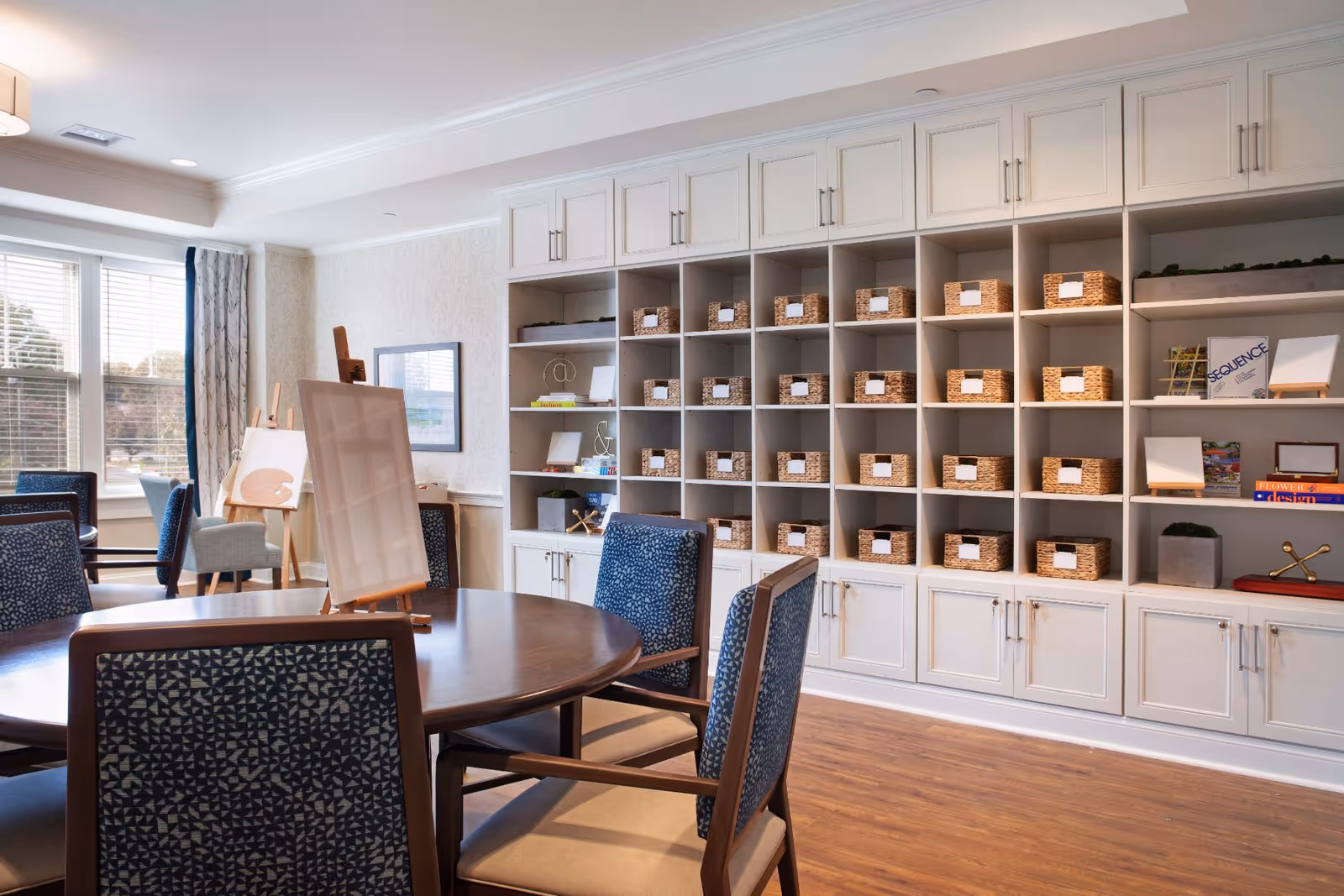 A bright and spacious activity room with a round wooden table surrounded by blue patterned chairs. The room features a large shelving unit filled with wicker baskets and some decorative items. There are easels with blank canvases near a window with white blinds and curtains, allowing natural light to fill the room. The floor is wooden, and the walls are light-colored with subtle wallpaper.