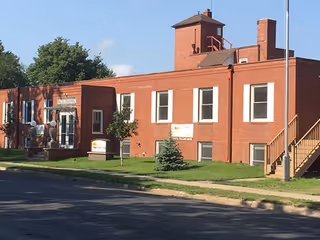 Front exterior of a one-story red brick assisted living building with a small tower, lawn, sidewalk, and exterior stairs.