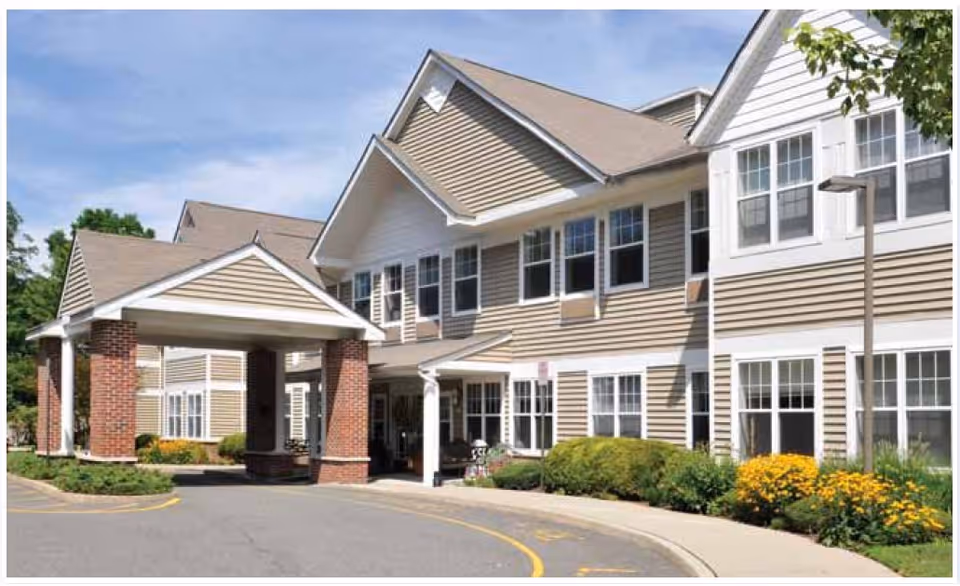 Exterior view of a senior living facility building with beige siding and white trim, featuring a covered entrance supported by brick columns, surrounded by landscaping with green bushes and yellow flowers under a blue sky.