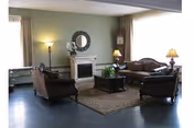 A cozy living room with a set of vintage-style sofas and armchairs arranged around a wooden coffee table on a patterned rug. The room features a small white fireplace with a round mirror above it, a tall floor lamp, a grandfather clock, and table lamps providing warm lighting. Large windows with curtains allow natural light to enter the space.