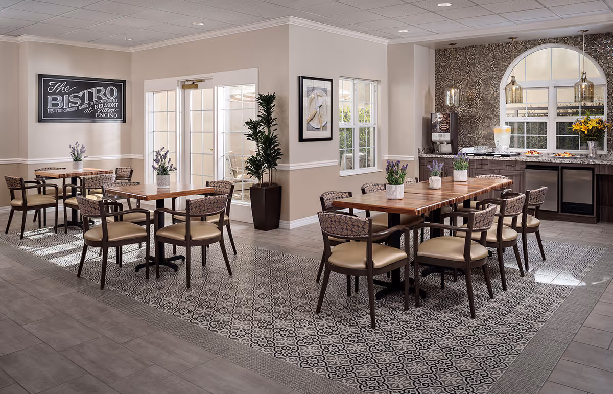 A bright and inviting dining area in a senior living facility with wooden tables and cushioned chairs arranged neatly on a patterned tile floor. The room features large windows, a decorative plant, and a buffet counter with a mosaic tile backsplash, pendant lights, and refreshments. A sign on the wall reads 'The Bistro at Belmont Village Encino.'
