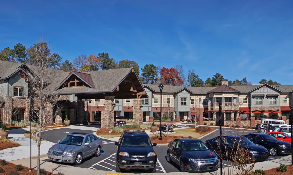 Exterior view of Village Park Peachtree Corners senior living facility showing a large two-story building with a covered entrance supported by stone pillars, a parking lot with several cars, landscaped grounds, and a clear blue sky.
