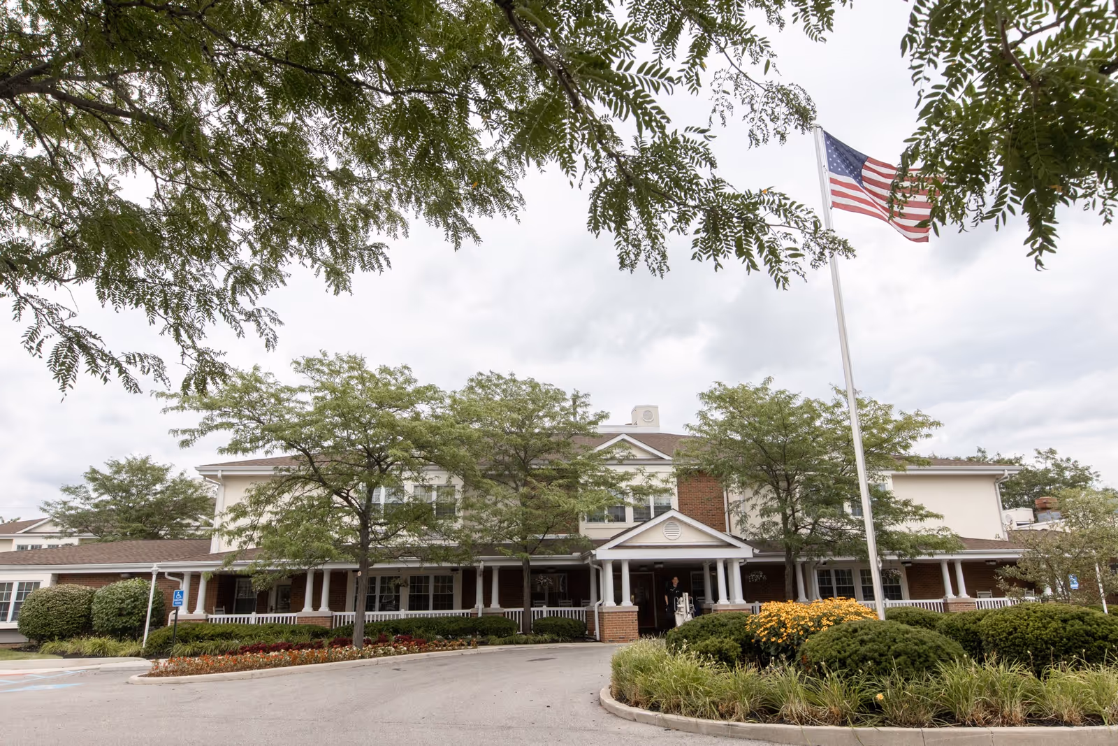 Front exterior view of Carriage Court Senior Living building with a covered entrance, surrounded by trees and landscaped bushes and flowers. An American flag is flying on a flagpole in front of the building under a cloudy sky.