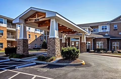 Entrance of a senior living facility with a covered drop-off area supported by stone and white pillars, surrounded by a multi-story building with windows and balconies under a clear blue sky.