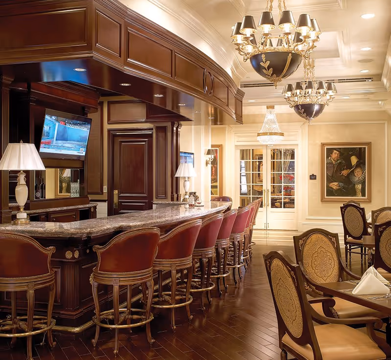 Elegant indoor dining and bar area with leather bar stools, a marble countertop, chandeliers, and upholstered dining chairs.