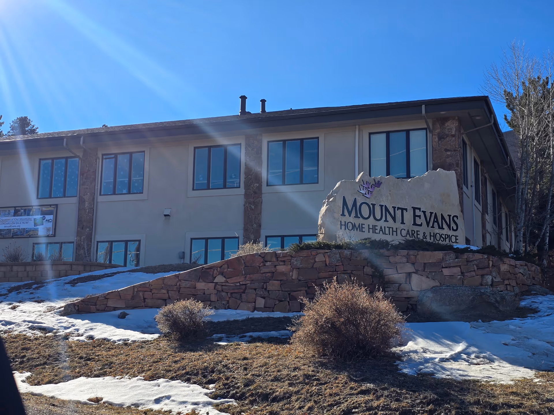 Exterior view of a two-story building with beige walls and stone accents under a clear blue sky. In front of the building, there is a large stone sign that reads 'Mount Evans Home Health Care & Hospice'. There is some snow on the ground and a few bushes near the sign.