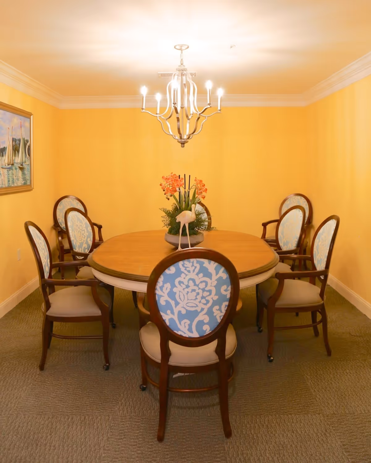 A dining room with a round wooden table surrounded by six upholstered chairs with blue and white patterned backs. A chandelier with lit bulbs hangs above the table, and a floral centerpiece decorates the table. The walls are painted yellow, and there is a painting of sailboats on the left wall.