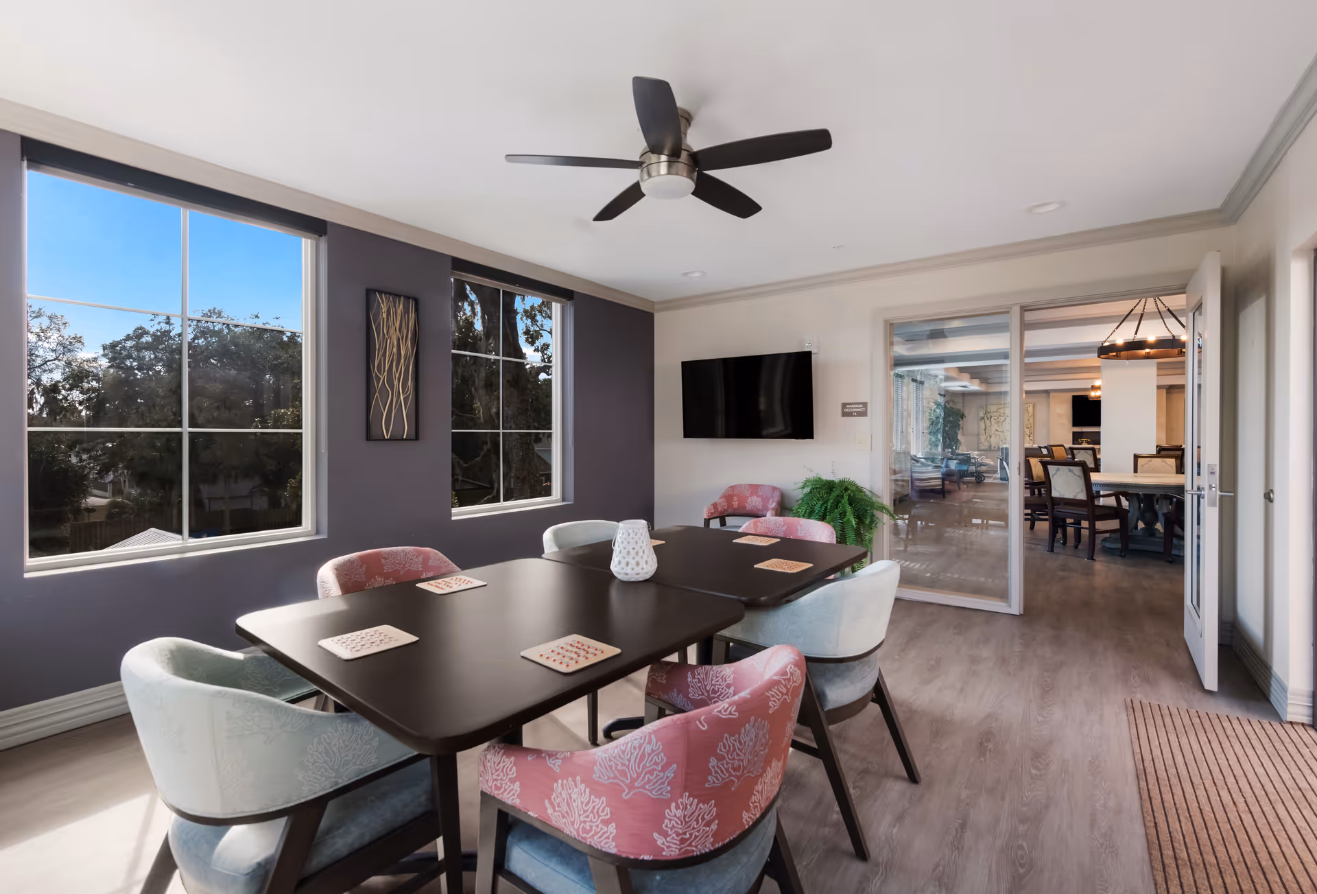 A bright and modern common area with a rectangular dark table surrounded by six upholstered chairs in pastel colors. The room has large windows with views of trees outside, a ceiling fan, a wall-mounted TV, and a sliding glass door leading to another room with more seating and a chandelier.