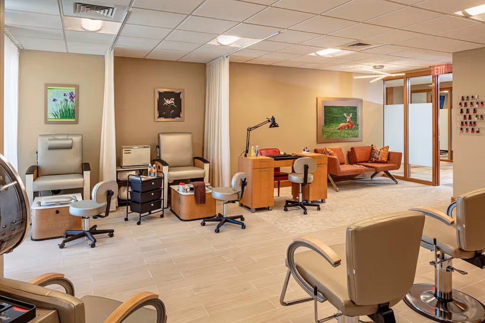 A well-lit salon area in a senior living facility with beige walls and tiled flooring. The room features pedicure chairs with foot baths, rolling stools, a manicure table with chairs, and a small seating area with an orange couch and decorative pillows. Artwork of flowers and a deer adorn the walls, and a nail polish display is visible near the entrance.
