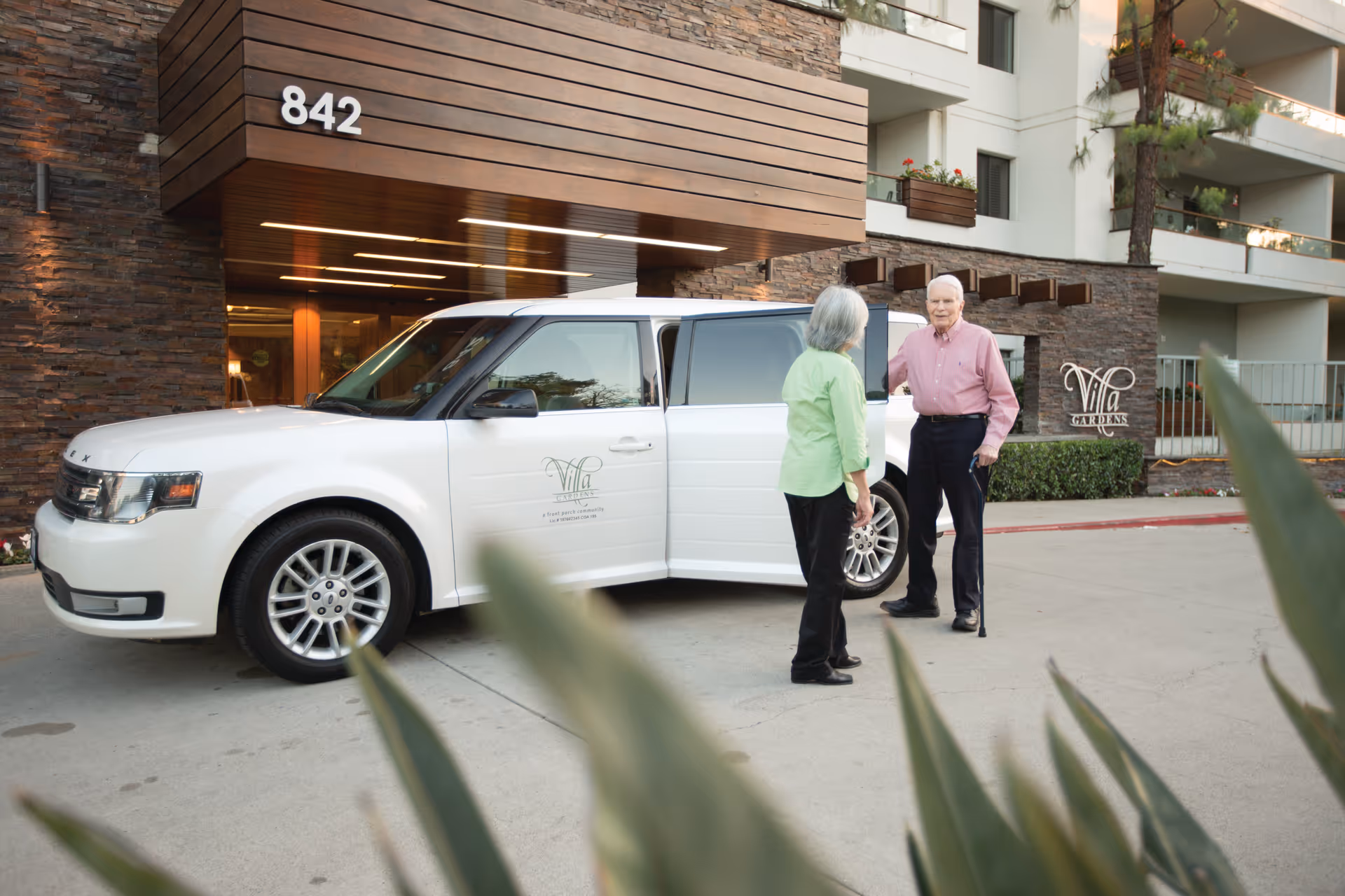 An elderly man with a cane and an elderly woman stand outside a building near a white vehicle with the Villa Gardens logo. The building has a modern exterior with stone and wood accents, and the number 842 is displayed above the entrance.