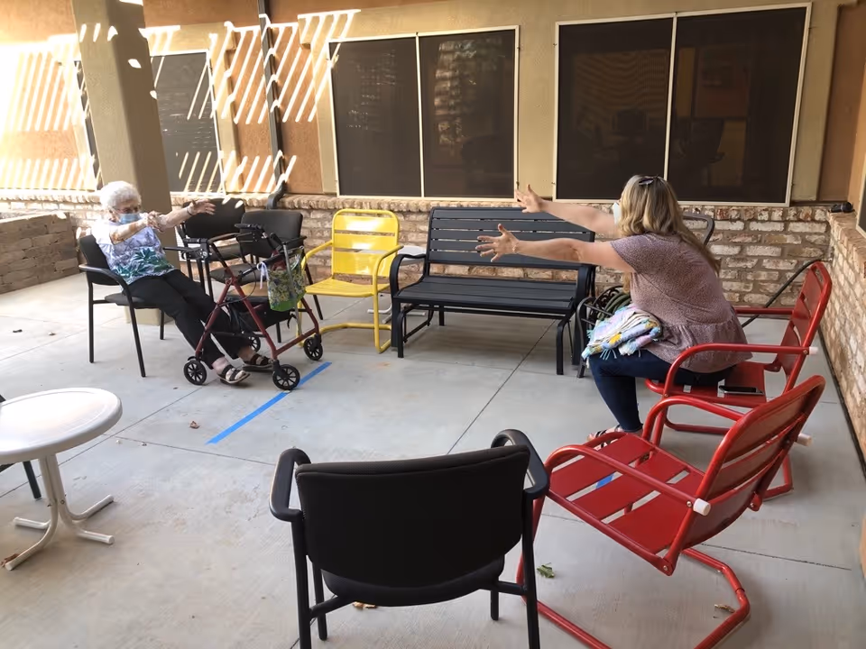 Two women sitting on chairs facing each other on a covered patio. One woman is elderly with a walker in front of her, and the other woman is reaching out towards her. The patio has various colored chairs and a small white table, with brick walls and windows in the background.