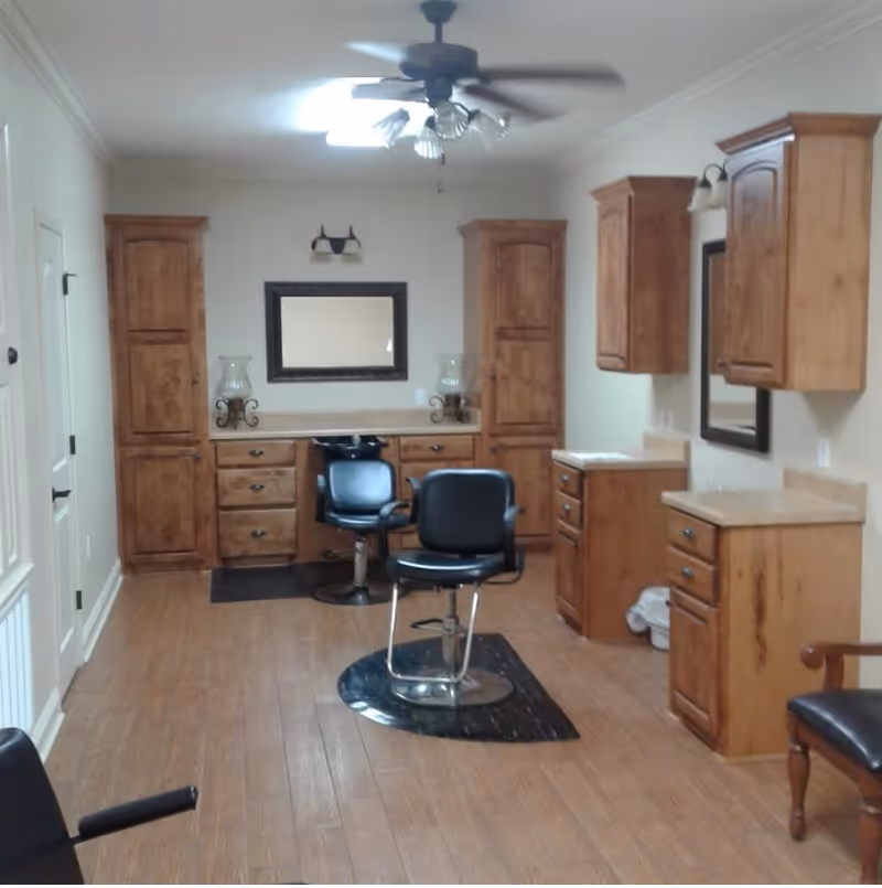 Interior salon-style room with two black salon chairs, wooden cabinets and mirrors, and a ceiling fan over a wood floor.