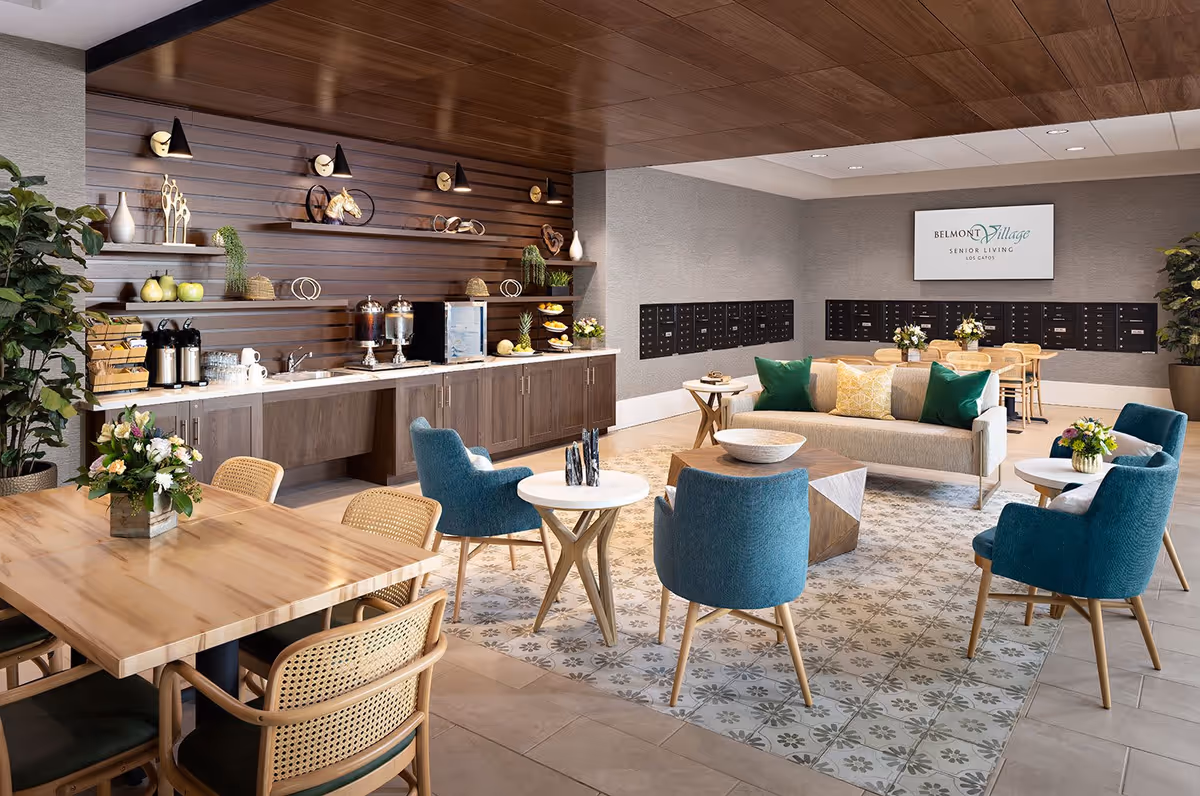A modern senior living common area with a wooden ceiling and a patterned tile floor. The space features a beige sofa with green and yellow pillows, four blue armchairs around small round tables, and a wooden dining table with wicker chairs. Along one wall, there is a countertop with cabinets, beverage dispensers, a mini fridge, and decorative shelves with plants and ornaments. A large screen on the wall displays the Belmont Village Senior Living Los Gatos logo. Mailboxes are visible on the back wall, and there are potted plants and floral arrangements throughout the room.