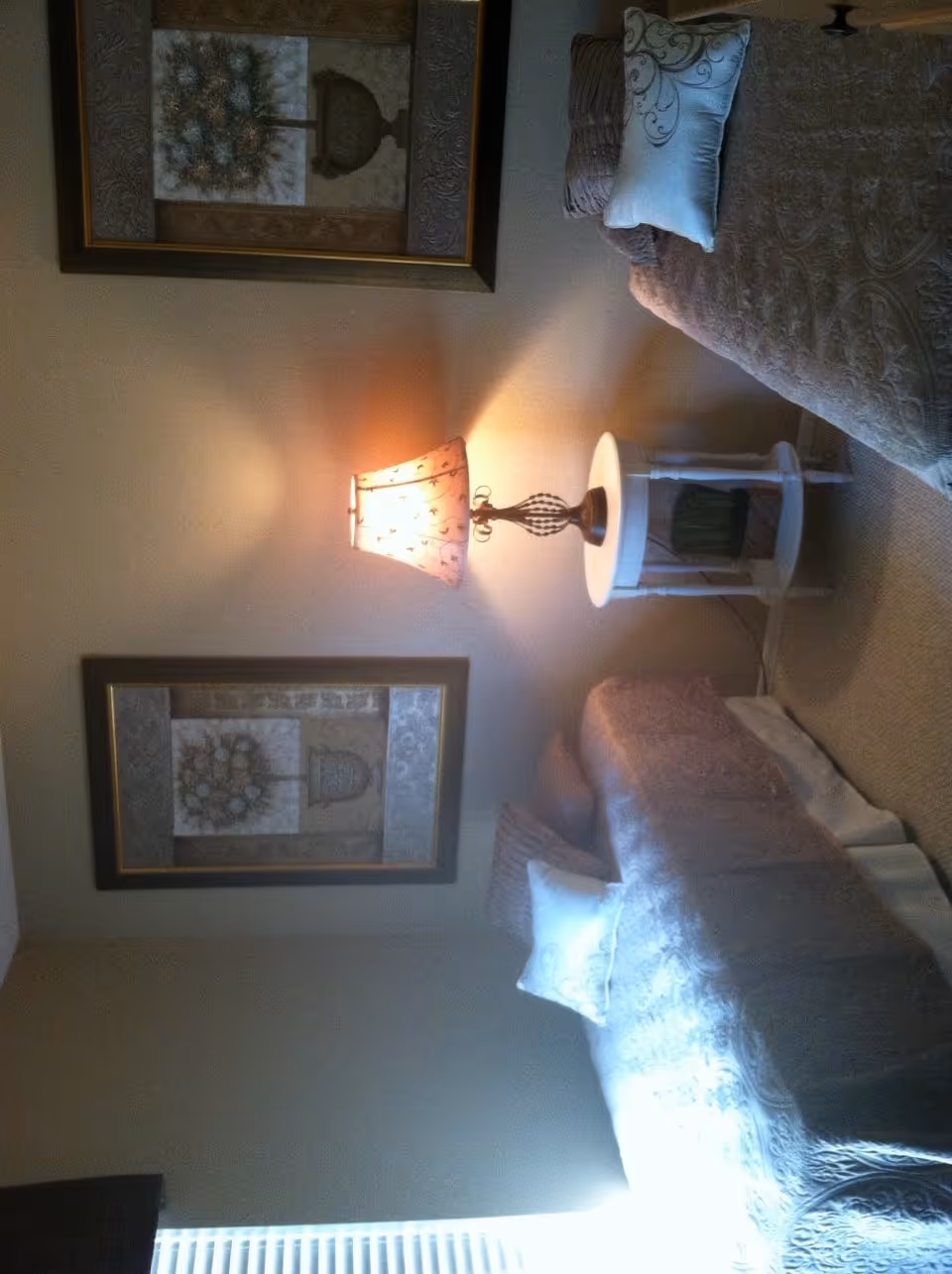 A cozy bedroom with two twin beds covered in patterned bedspreads and decorative pillows. Between the beds is a small round white nightstand with a decorative lamp emitting warm light. Two framed floral artworks hang on the beige wall above the nightstand. A window with blinds is partially visible on the left side of the image.