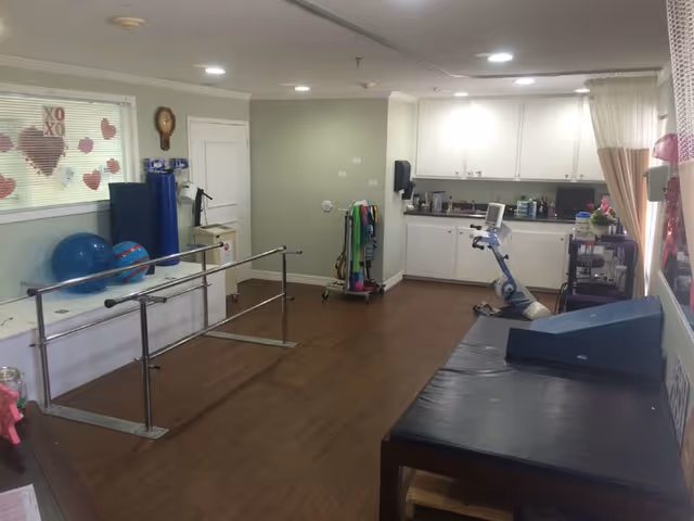 A rehabilitation room with wooden flooring featuring parallel bars for walking exercises, exercise balls, resistance bands, a stationary exercise bike, and a padded therapy table. The room has white cabinets, a clock on the wall, and windows with heart-shaped decorations.
