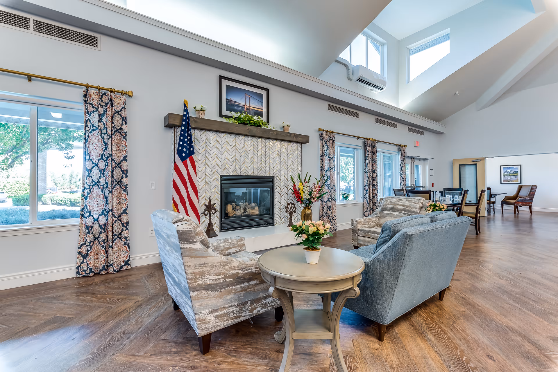 Bright, spacious senior living common area with seating around a fireplace, decorative curtains, an American flag, and skylights.