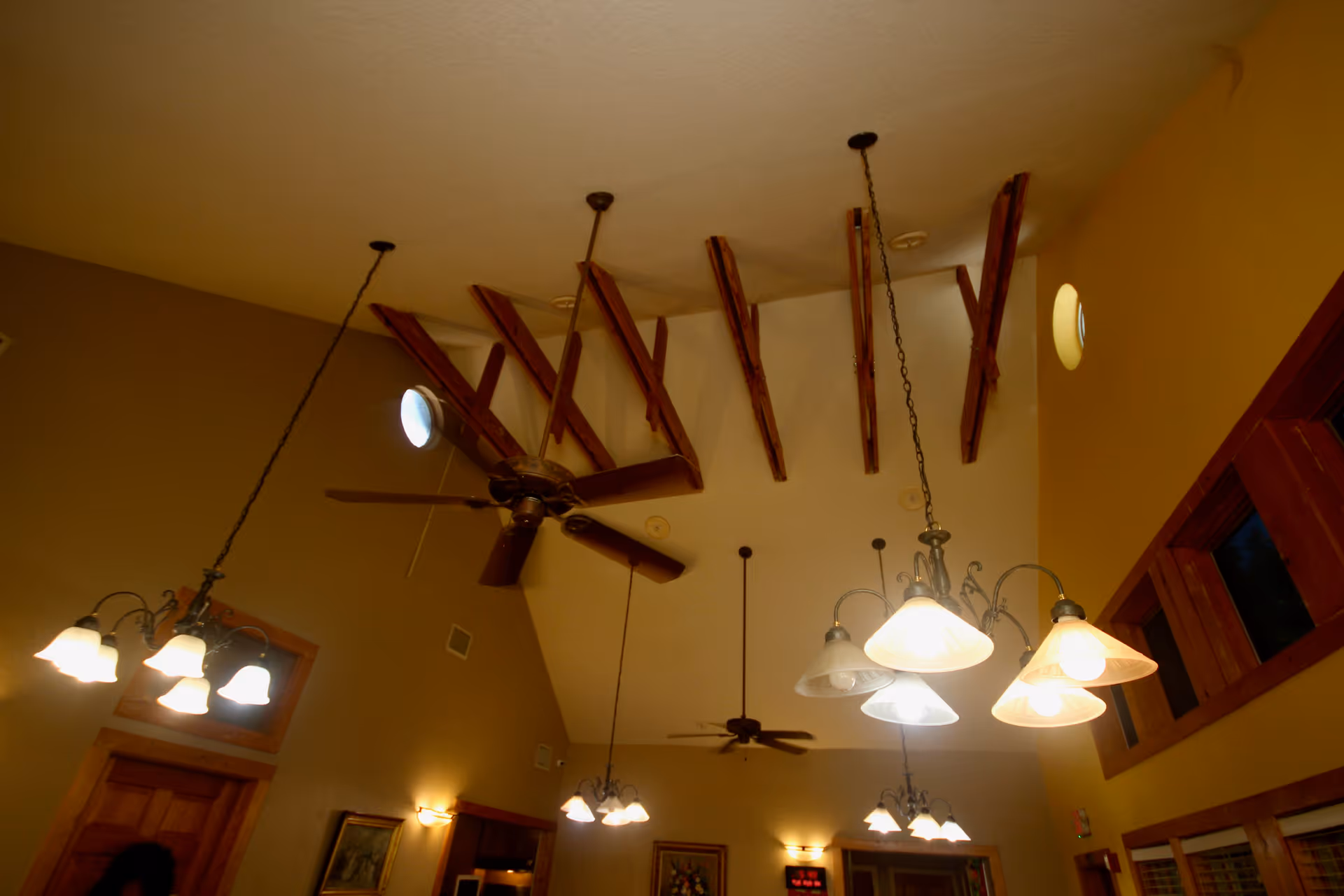 Interior view of a room with a high vaulted ceiling featuring exposed wooden beams, ceiling fans, and multiple hanging light fixtures with frosted glass shades. The walls are painted in warm tones with wooden window frames and a wooden door visible.