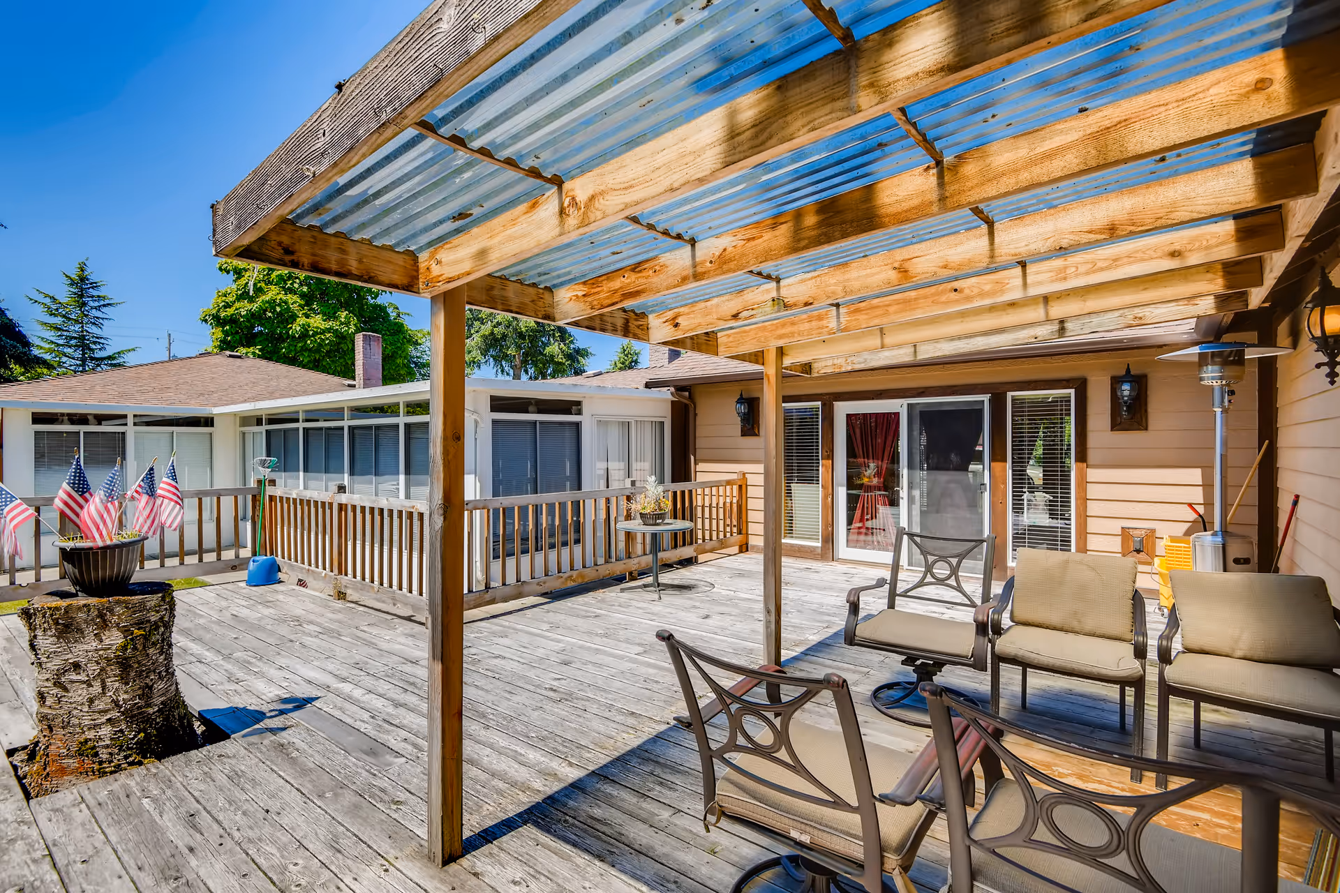 Outdoor wooden deck area with a pergola covered by a translucent roof. The deck has several cushioned chairs and a small table. There is a planter with small American flags on a tree stump, and the background shows parts of the building with windows and doors under a clear blue sky.