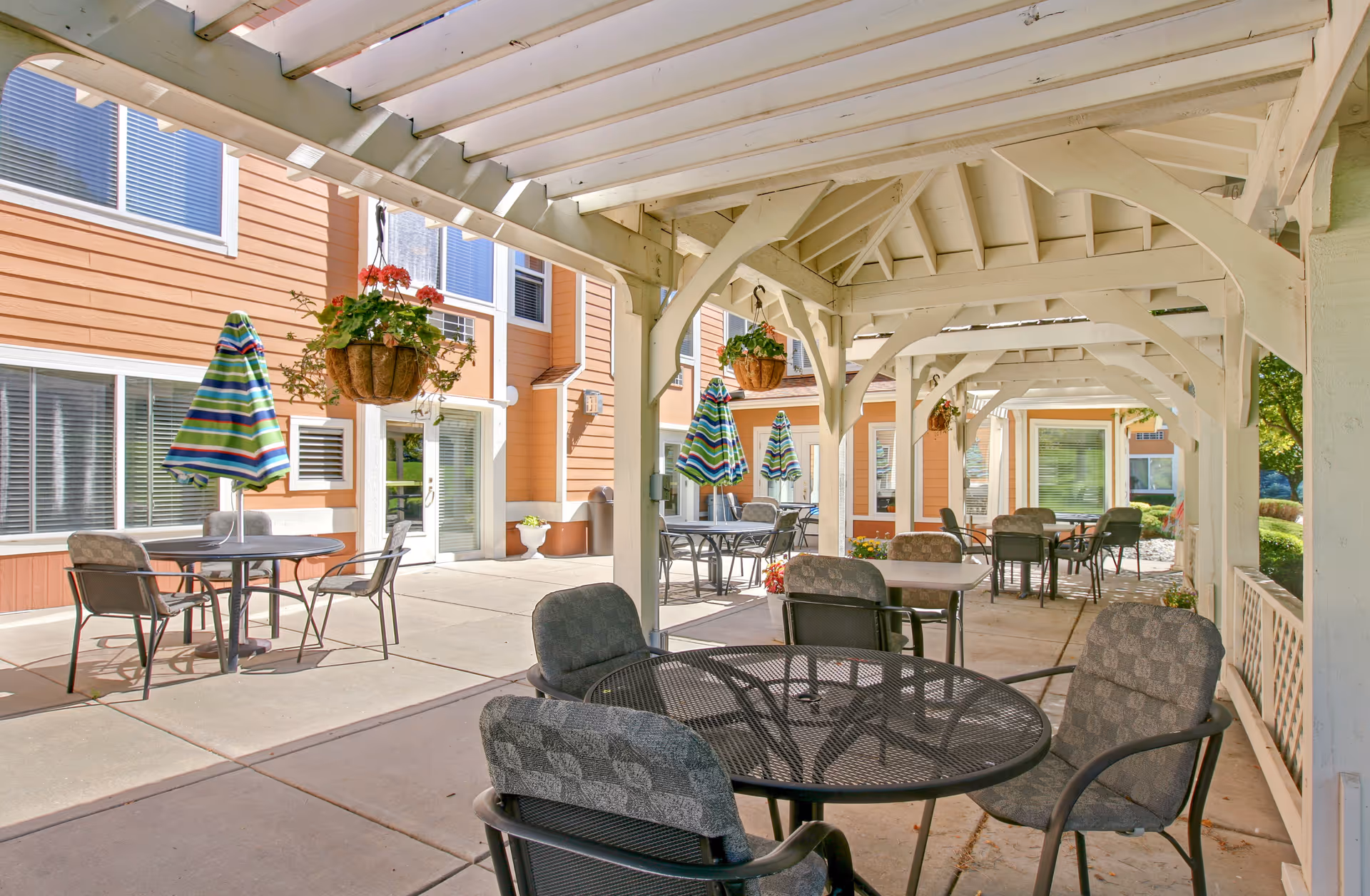 Outdoor covered patio area with multiple round tables and cushioned chairs. Some tables have closed striped umbrellas. Hanging flower baskets are suspended from the white wooden beams of the patio roof. The patio is adjacent to a peach-colored building with white trim and several windows.