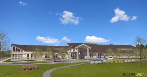Exterior view of a single-story senior living facility building with a brown roof and beige walls, surrounded by green grass, small trees, and a few parked cars under a blue sky with scattered clouds.